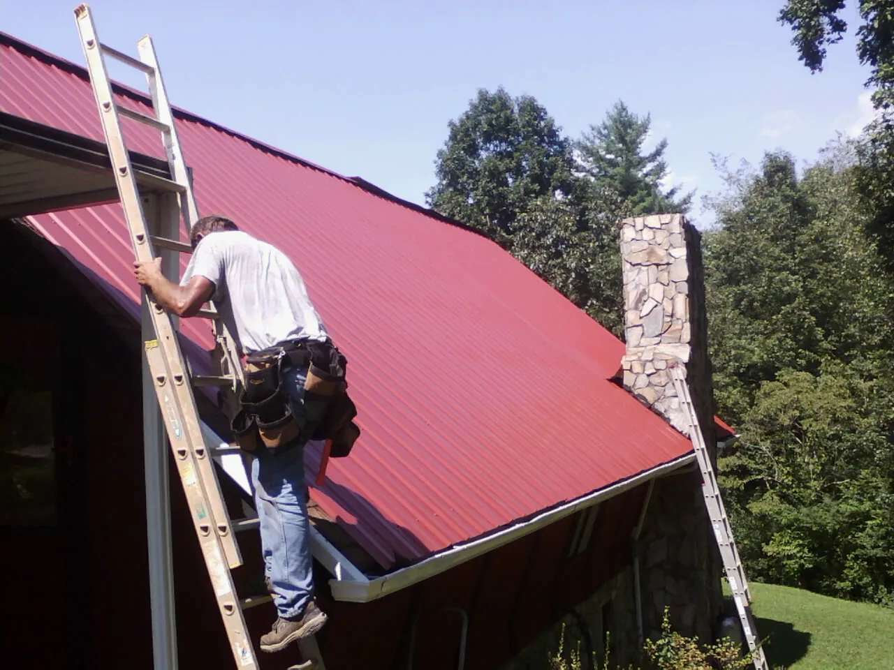 Roofer on a ladder beside a red metal roof and stone chimney — Soffit & Fascia Repair by Roofing Brothers in Hilltown